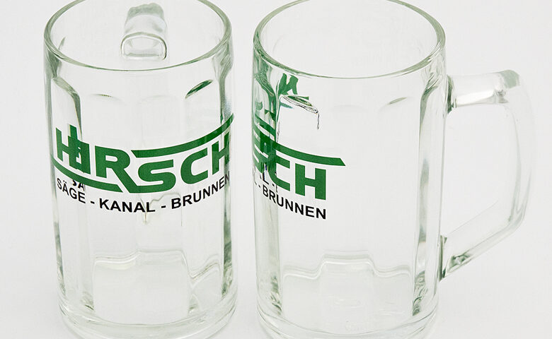 Zwei leere gläserne Bierkrüge mit Henkel bedruckt mit dem grüen Schriftzug Hirsch auf hellem Hintergrund
