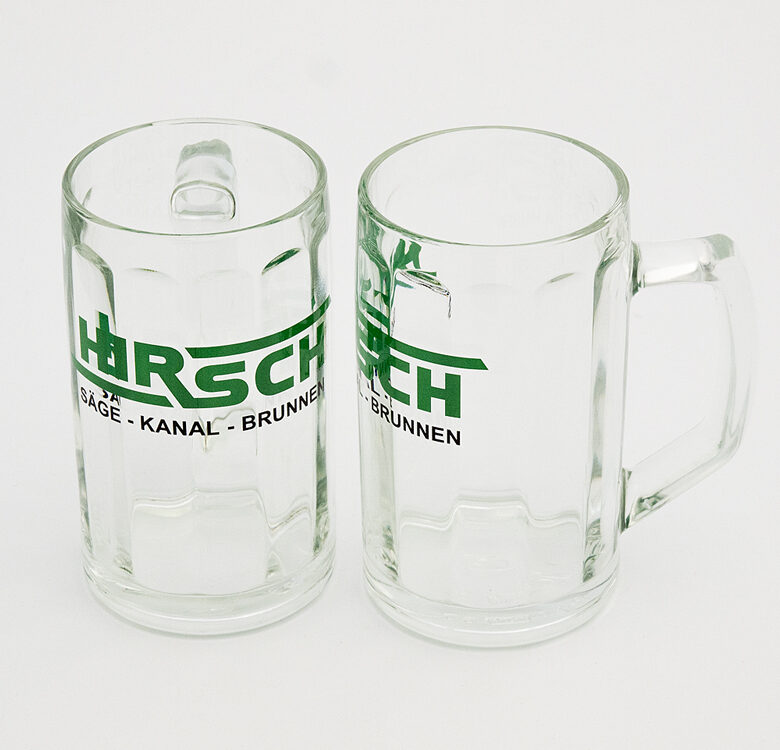 Zwei leere gläserne Bierkrüge mit Henkel bedruckt mit dem grüen Schriftzug Hirsch auf hellem Hintergrund