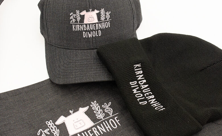 graue Kappe, schwarze Wollhaube und graue Schürze mit dem Logo in weiß und rosarot der Fa. Kirnbauernhof Diwold bestickt