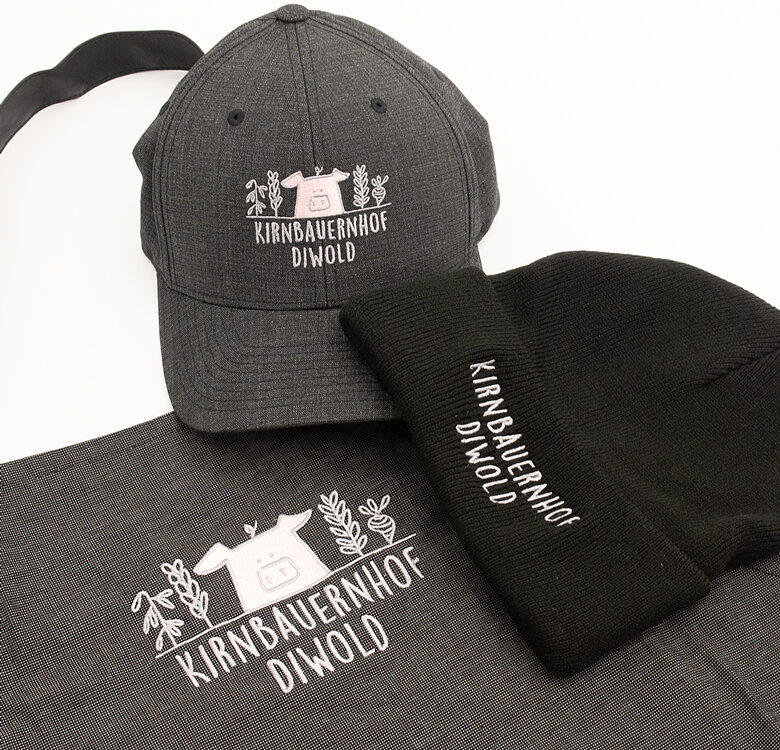 graue Kappe, schwarze Wollhaube und graue Schürze mit dem Logo in weiß und rosarot der Fa. Kirnbauernhof Diwold bestickt